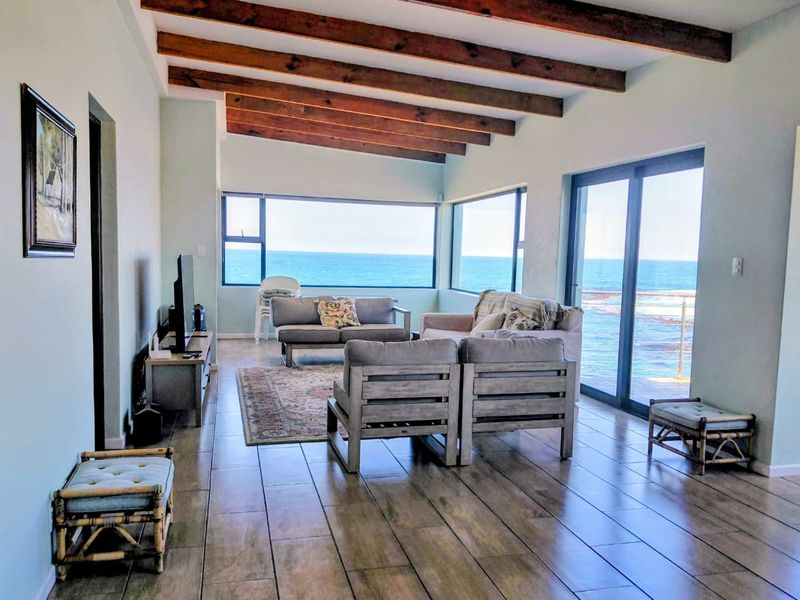 Spacious living area featuring exposed timber ceiling beams, tiled flooring, and expansive windows offering panoramic ocean views with direct access to the outdoors.