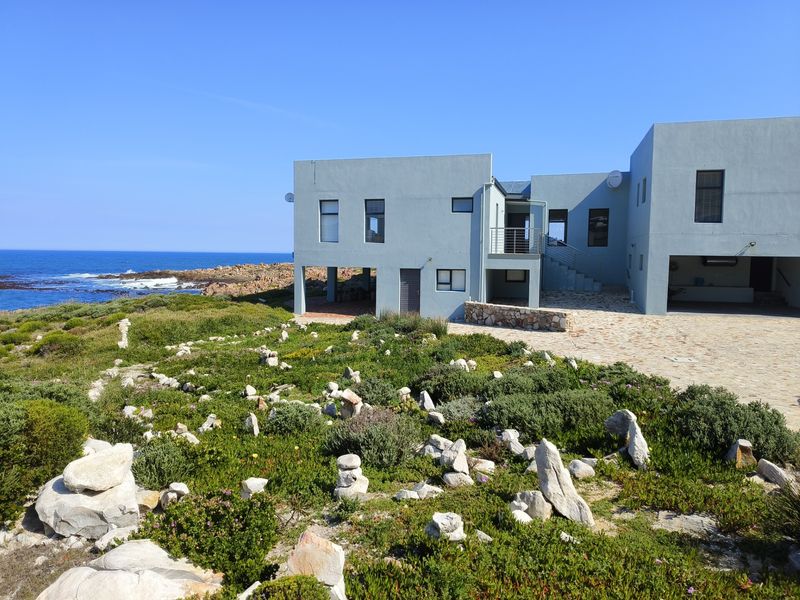 A modern coastal home featuring a stone-paved driveway, external staircase, and expansive ocean views, set within a natural landscaped environment.
