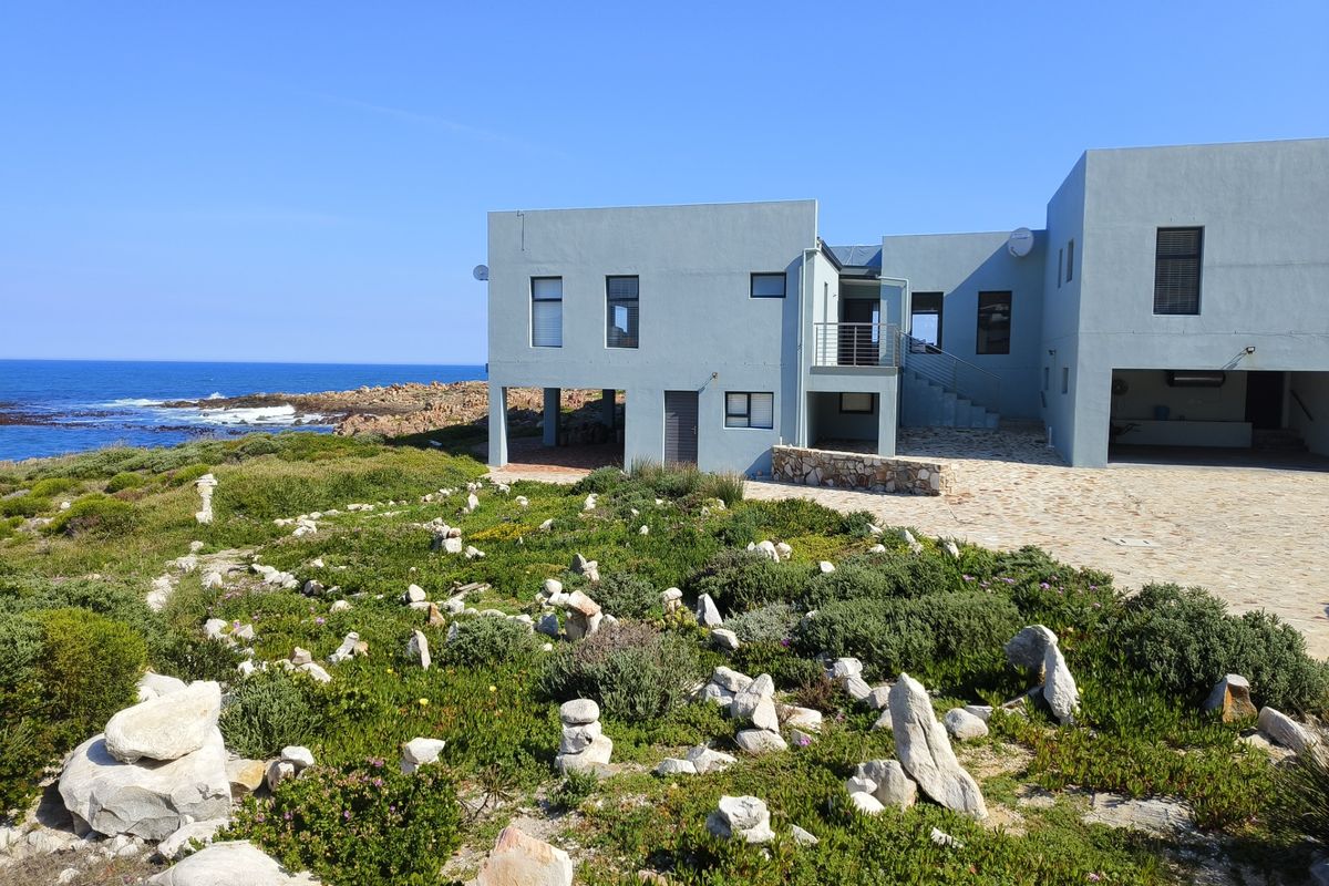 A modern coastal home featuring a stone-paved driveway, external staircase, and expansive ocean views, set within a natural landscaped environment.