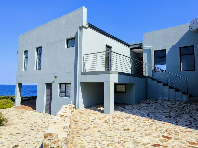 Rear elevation of a modern coastal home featuring stone paving, an elevated balcony with stainless steel railings, and an external staircase.