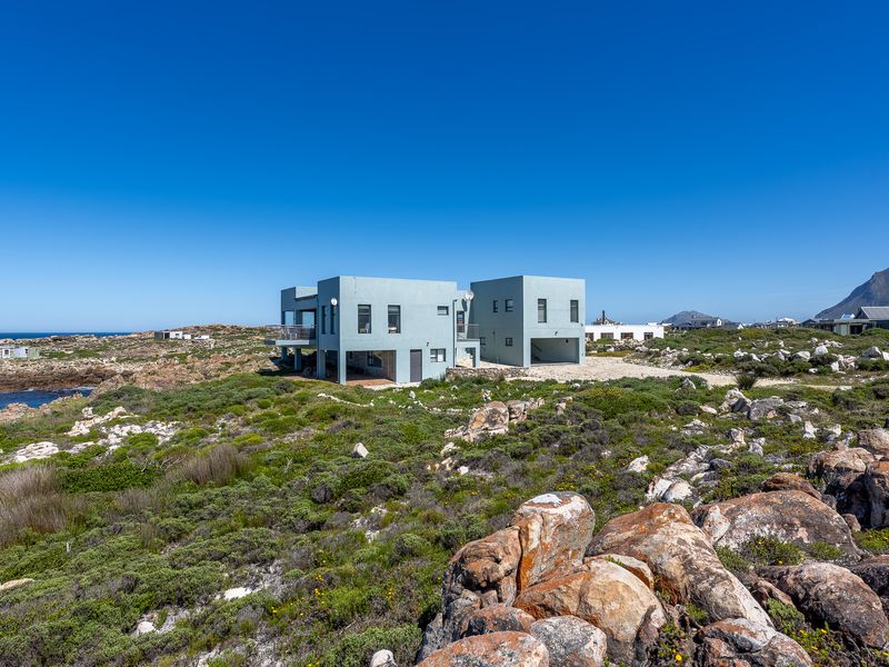 A modern, multi-level coastal home featuring a balcony and integrated garage, situated in a rugged, rocky landscape overlooking the ocean.