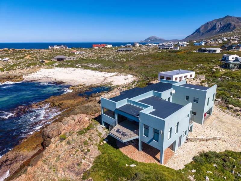 Aerial view of a modern coastal residence situated on a rocky shoreline, featuring a flat roof design and an elevated balcony overlooking the ocean.