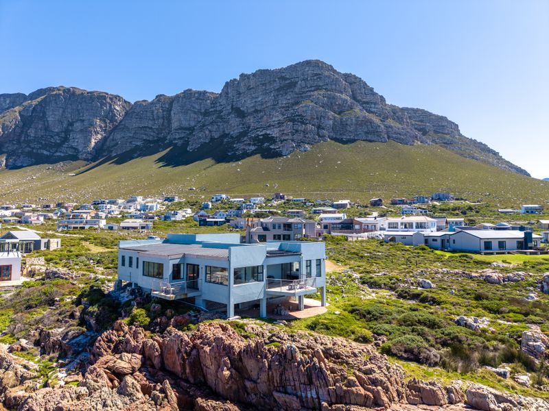 Aerial view of a modern coastal residence featuring a flat roof and balconies, set against a dramatic mountain backdrop.