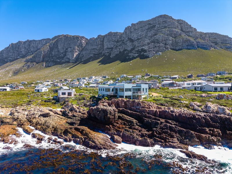An aerial view of a modern coastal residence situated on a rocky shoreline with dramatic mountain backdrop and ocean views.