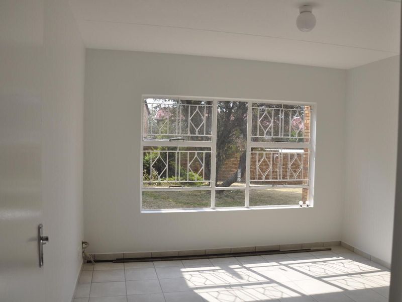 A bright, empty bedroom featuring neutral tiled flooring, white walls, and a large window fitted with security burglar bars.