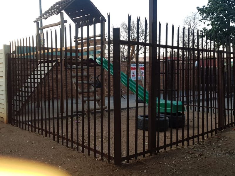 A fenced outdoor play area featuring a brick boundary wall and paved ground, secured by metal perimeter fencing.