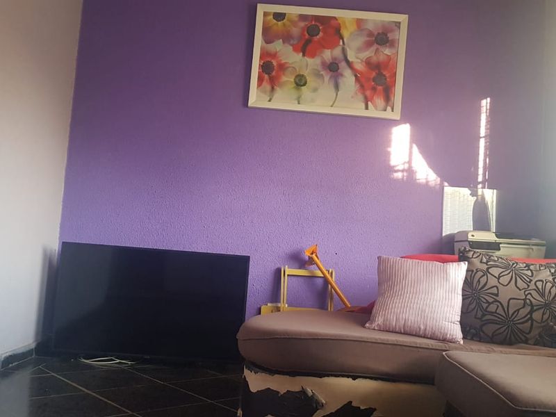 A living room area featuring tiled flooring and a purple accent wall, currently furnished with a sofa and television.