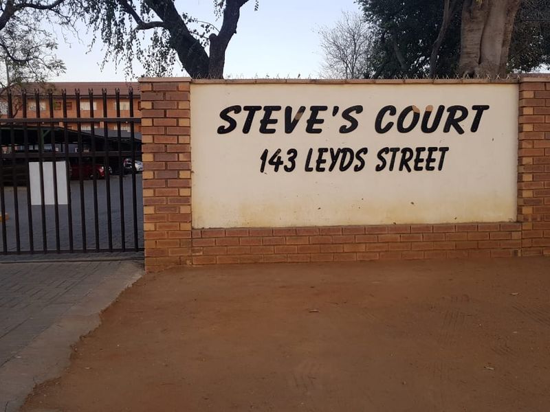 Front entrance view of Steve's Court apartment complex, featuring a brick boundary wall, metal security gate, and paved driveway access.