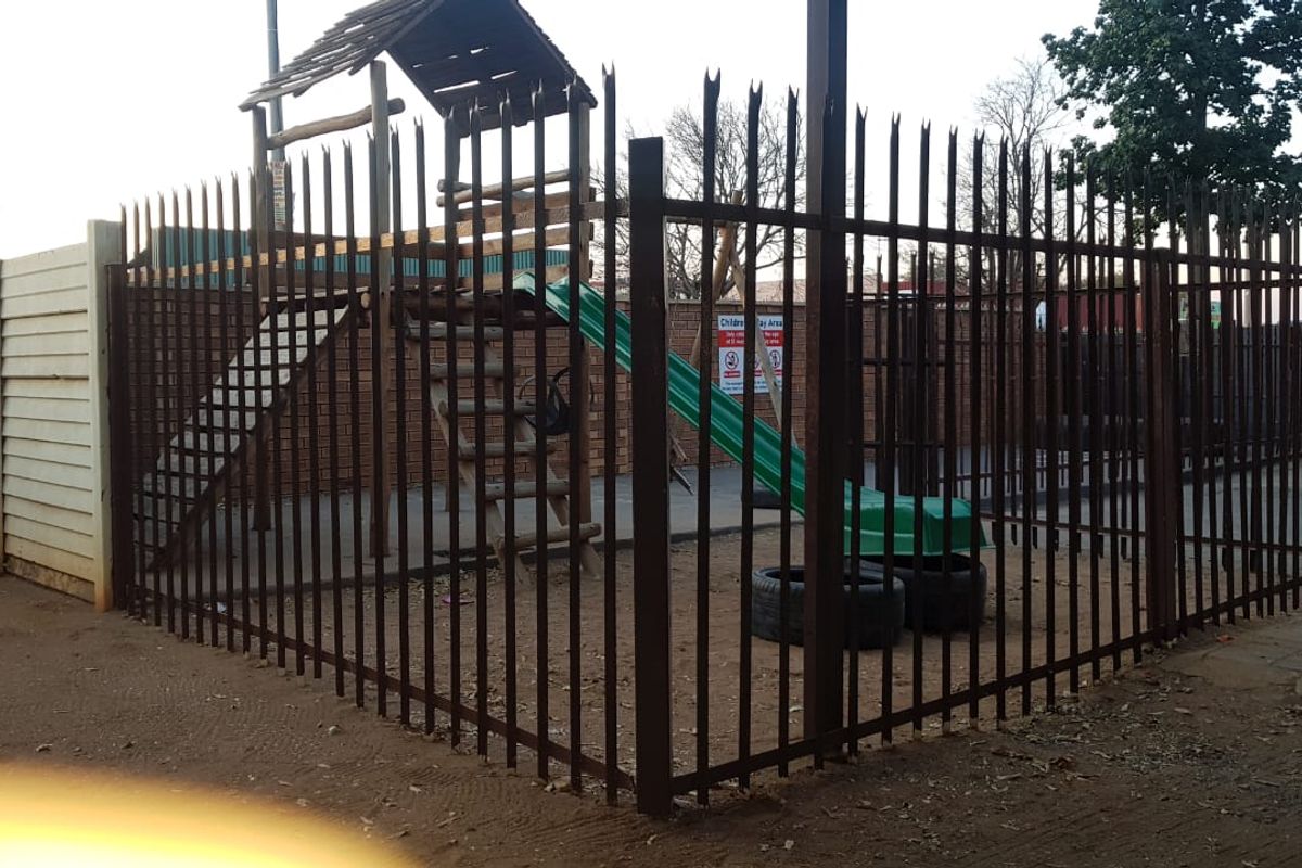 A fenced outdoor play area featuring a brick boundary wall and paved ground, secured by metal perimeter fencing.
