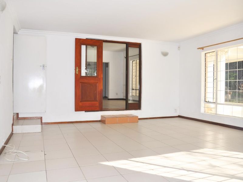 A bright, empty living room featuring tiled flooring, large windows with security bars, and a wooden door leading to an adjacent space.
