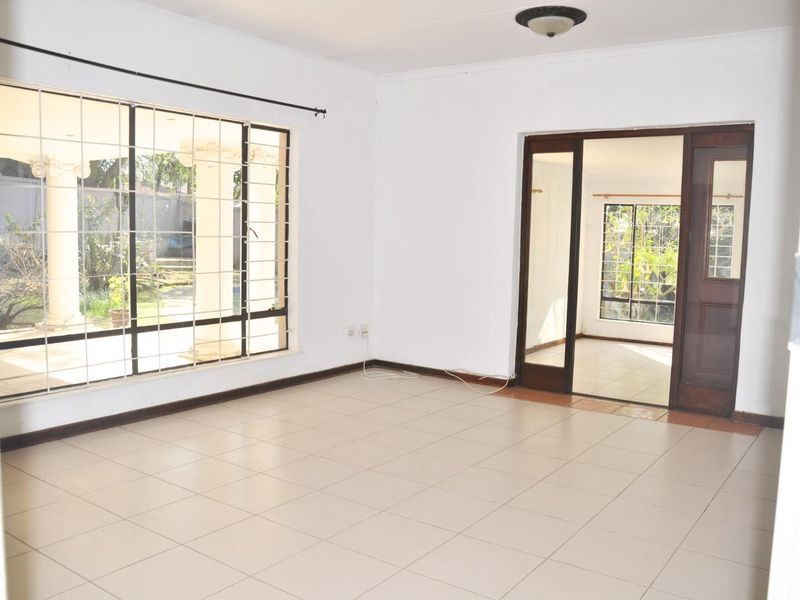 A bright, empty living room featuring neutral floor tiles, large windows with security bars, and a wooden door leading to an adjacent room.