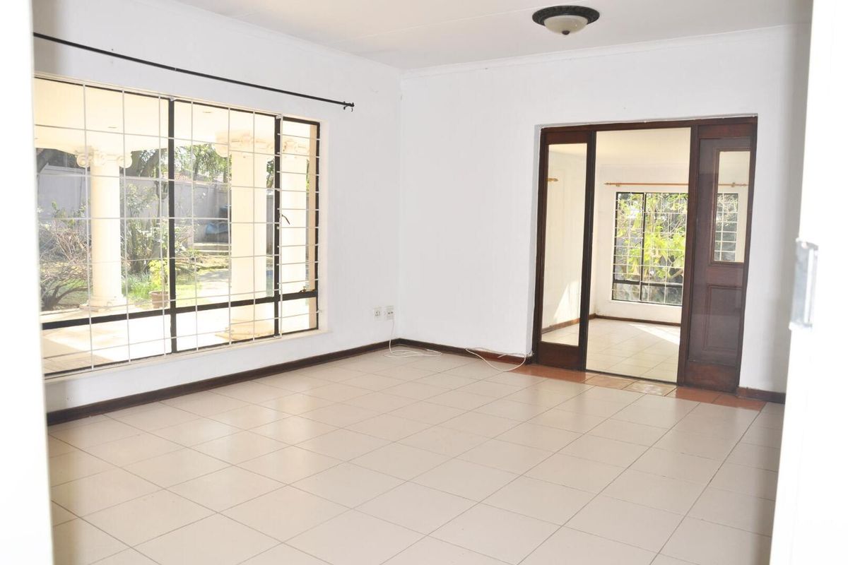 A bright, empty living room featuring neutral floor tiles, large windows with security bars, and a wooden door leading to an adjacent room.