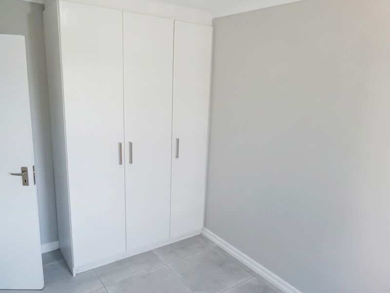 A clean, empty bedroom featuring neutral grey walls, modern tiled flooring, and a white built-in wardrobe with sleek handles.