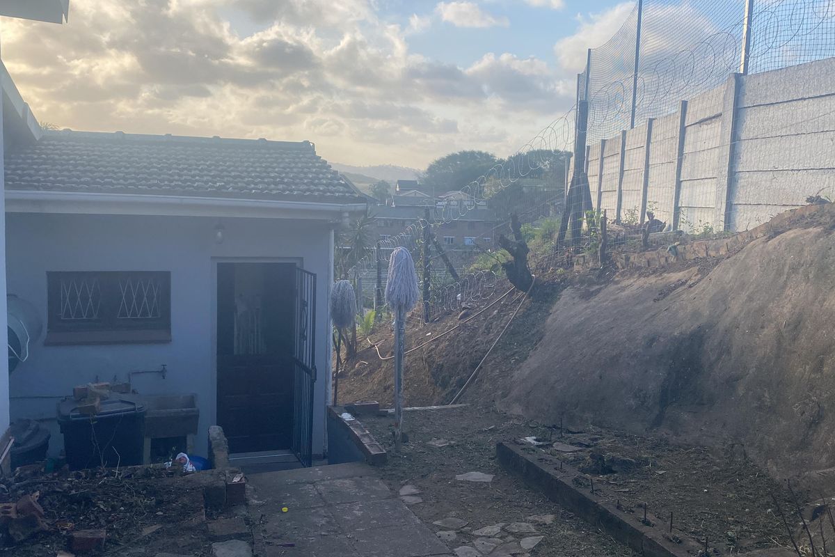 Rear exterior view of the property showing a utility entrance, concrete walling with security fencing, and an unlandscaped sloped garden area.