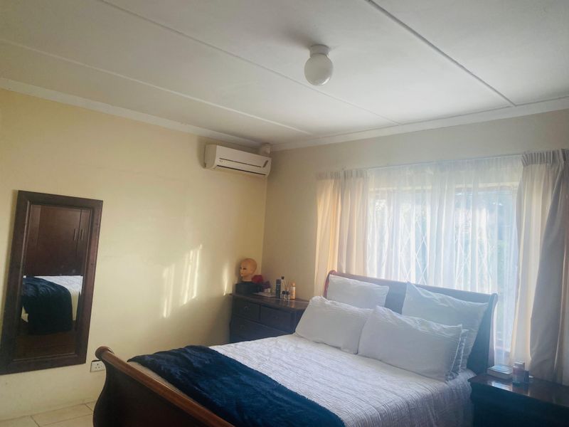 A functional bedroom featuring tiled flooring, a wall-mounted air conditioning unit, and a window fitted with a security gate.