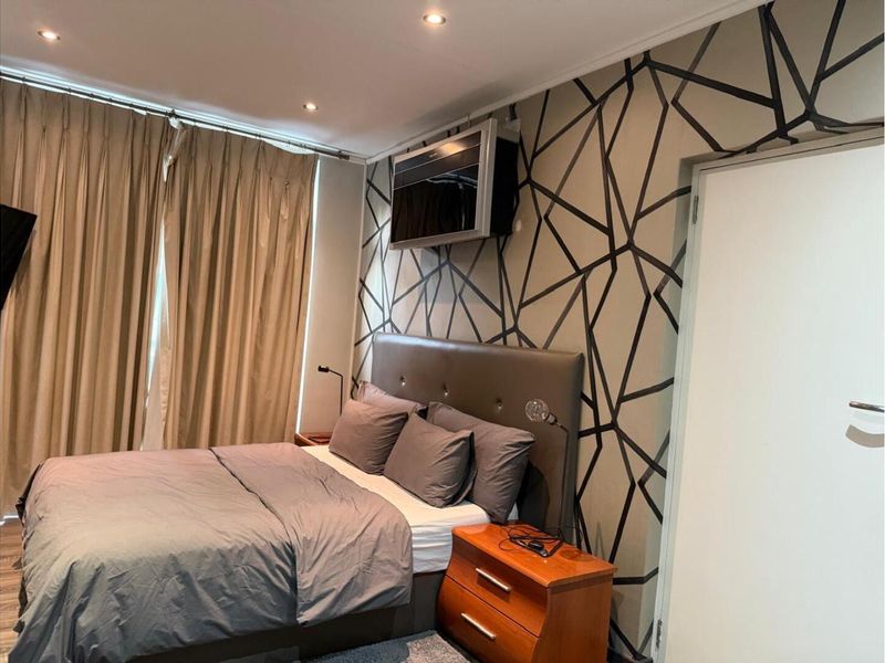 A well-maintained bedroom featuring recessed ceiling lighting, patterned feature wallpaper, and floor-to-ceiling curtains.