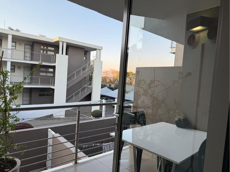 A covered balcony featuring stainless steel railings and tiled flooring, offering an elevated view of the apartment complex and surrounding grounds.