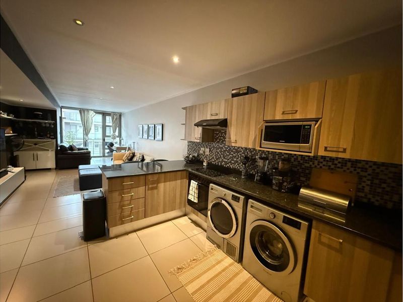 Open-plan apartment featuring a kitchen with wood-finish cabinetry, granite countertops, mosaic backsplash, and integrated laundry appliances, flowing into a living area with balcony access.