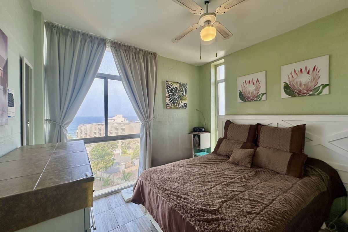 A bedroom featuring tiled flooring, a ceiling fan, and a large window offering coastal views, with painted brick walls.