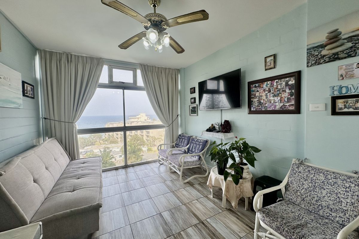 A bright living room featuring tiled flooring, a ceiling fan, and large windows offering scenic coastal views.