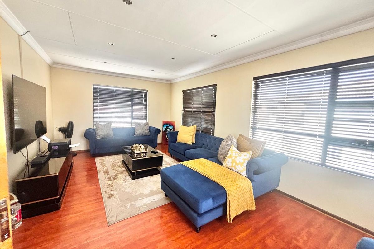 A spacious living room featuring warm-toned laminate flooring, recessed ceiling lighting, and large windows fitted with dark horizontal blinds.