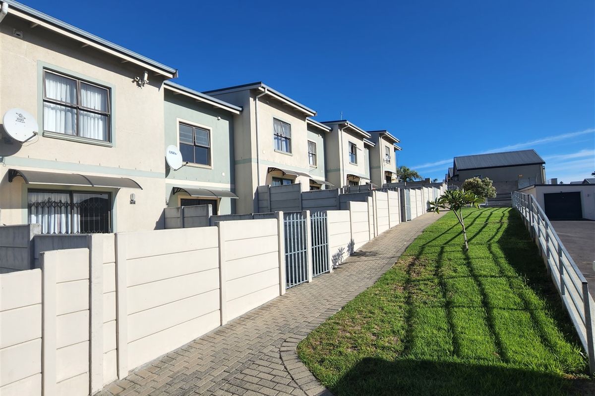 Rear view of a townhouse complex featuring a paved walkway, manicured lawn, and individual unit access with security gates and boundary walls.