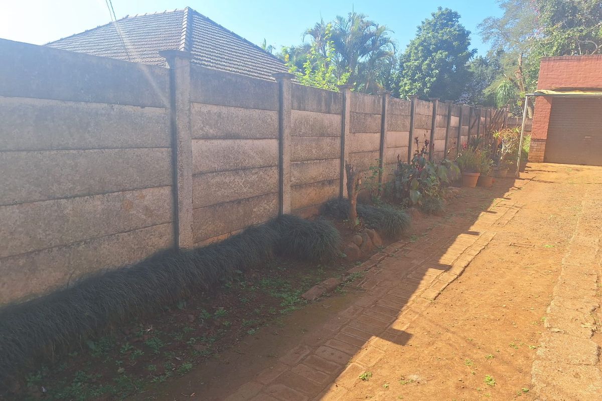 A side exterior view showing a long precast concrete boundary wall, unpaved ground, and basic landscaping along the property perimeter.