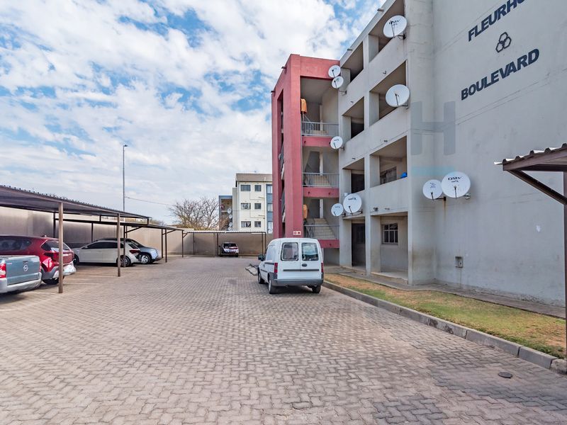 Exterior view of the apartment complex featuring a paved parking area with covered bays and a multi-story building facade.