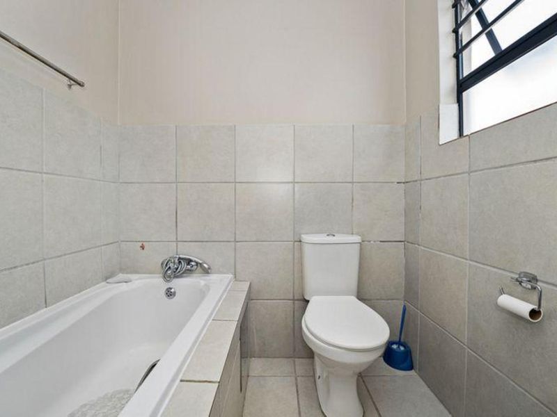 A functional bathroom featuring a built-in bathtub, a standard toilet, and neutral wall tiling with a security-barred window.