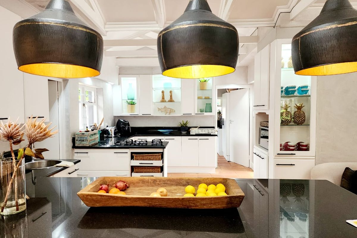 Modern kitchen featuring white cabinetry, black granite countertops, integrated lighting, and a central island with pendant lighting under a decorative beamed ceiling.