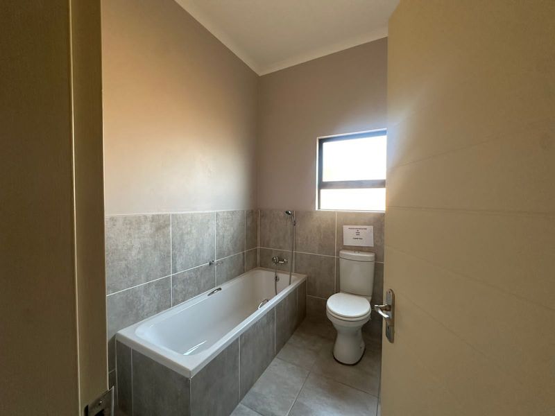 A clean, modern bathroom featuring a built-in bathtub with shower mixer and a standard toilet, finished with neutral grey wall and floor tiles.