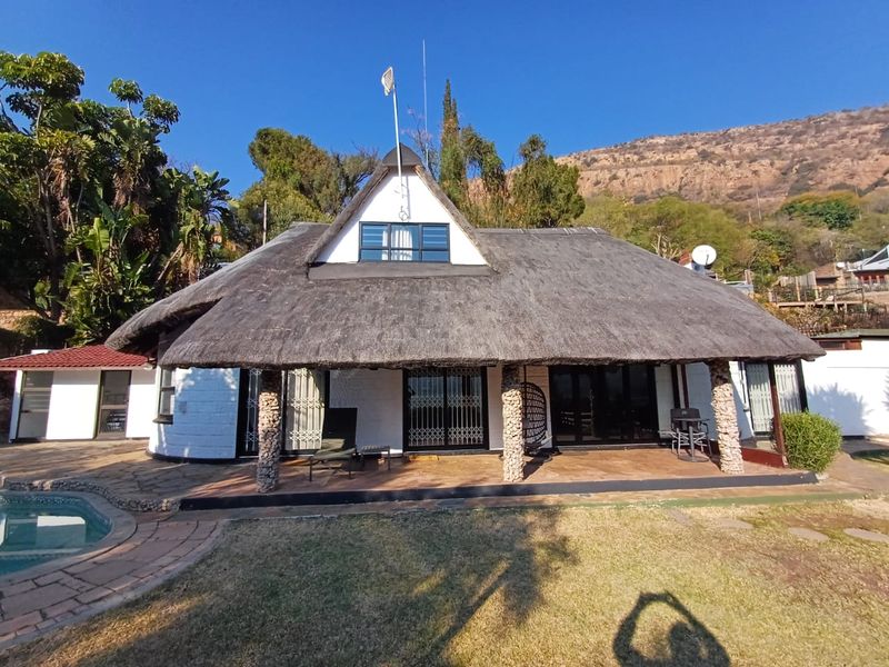 Rear view of a thatched-roof house featuring a covered patio, stone-clad pillars, and an adjacent swimming pool set against a mountain backdrop.