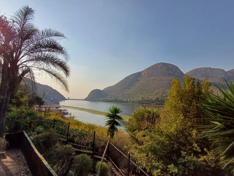 Scenic elevated view overlooking a tranquil lake and surrounding mountains, featuring established garden landscaping and secure perimeter fencing.