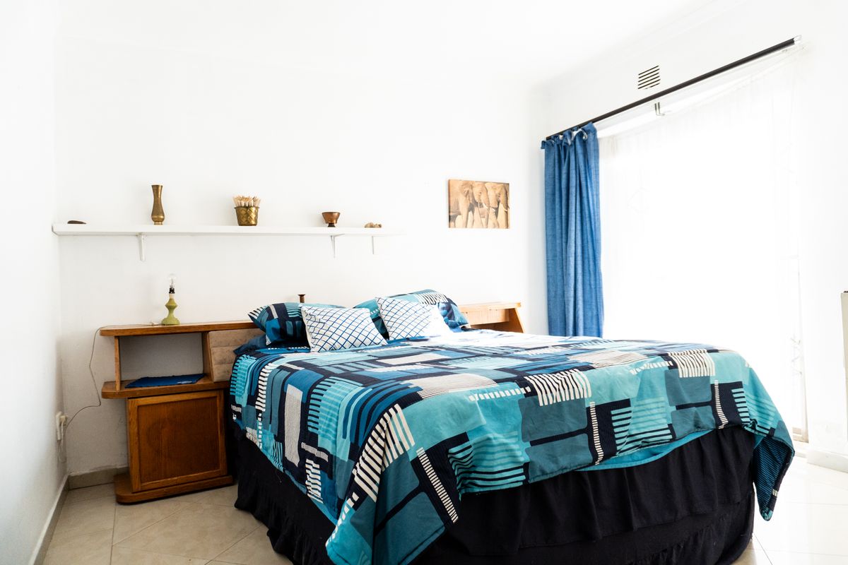 A bedroom featuring tiled flooring, a built-in wooden headboard with integrated shelving, and a large window with curtain rail.