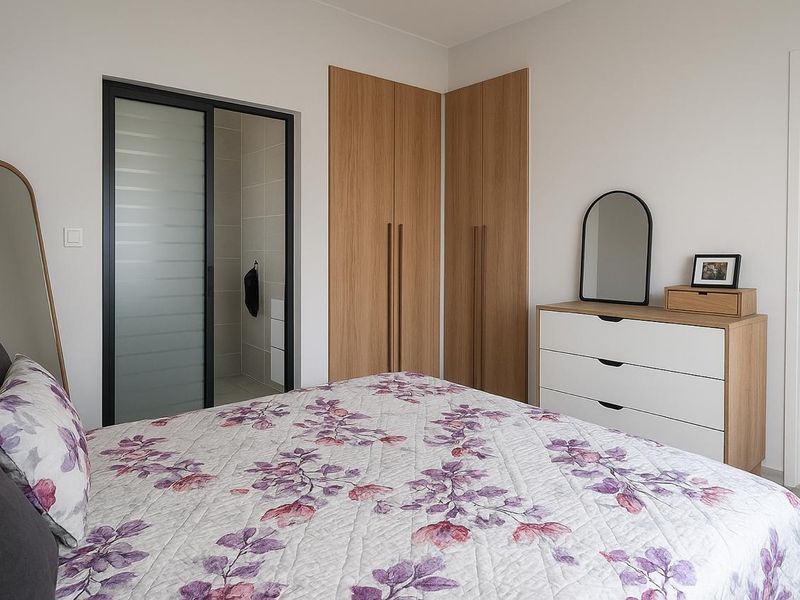 A modern bedroom featuring light wood built-in wardrobes, tiled flooring, and a sliding glass door leading to an en-suite bathroom.