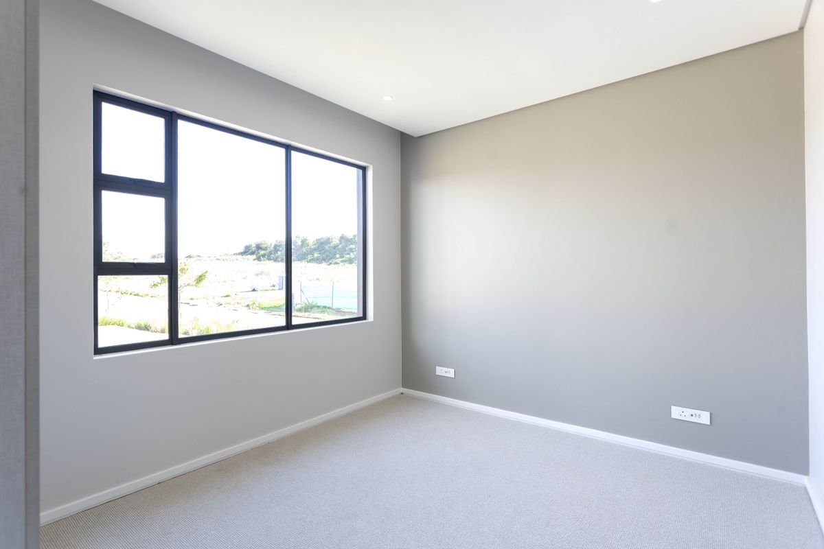 A bright, empty bedroom featuring neutral grey walls, plush carpeting, and a large window providing natural light and exterior views.