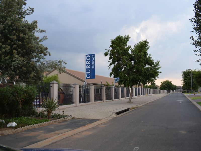 A street-level view of the property boundary featuring paved sidewalks, metal perimeter fencing, and established landscaping along the road.