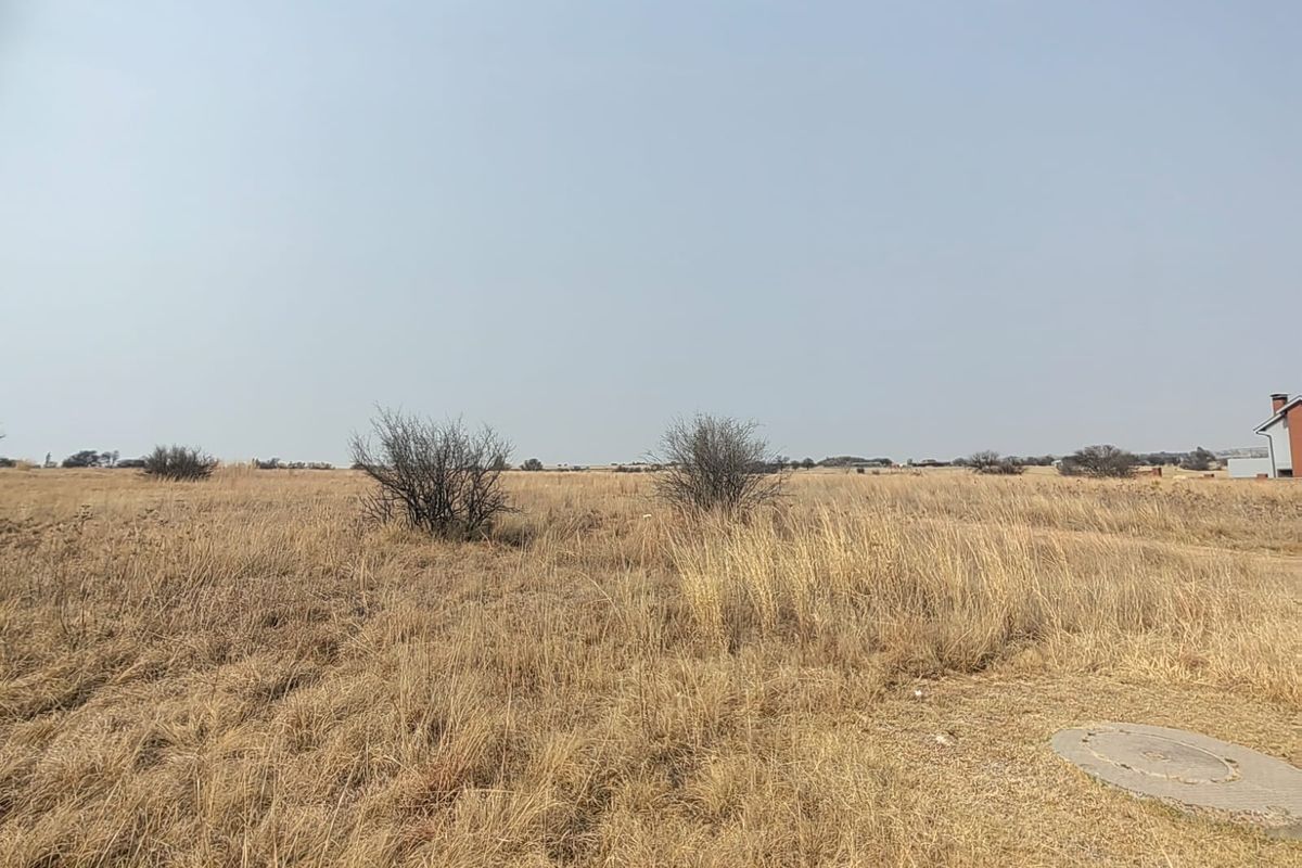 A wide-angle view of a vacant residential plot featuring dry grass, native shrubs, and a visible utility manhole cover.
