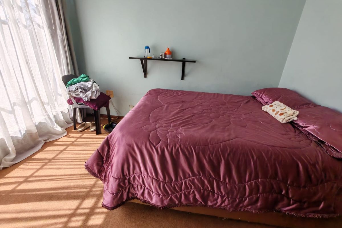 A bedroom featuring carpeted flooring and a wall-mounted shelf, with natural light entering through a large window.