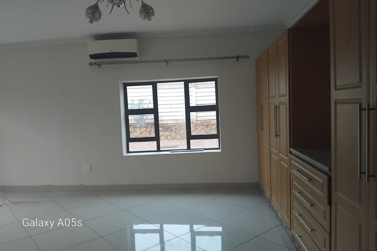 A bright bedroom featuring tiled flooring, built-in wooden cabinetry, a wall-mounted air conditioning unit, and a window with security bars.