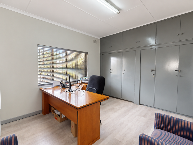 A functional home office featuring extensive built-in storage cupboards, light-toned laminate flooring, and a window with fitted venetian blinds.