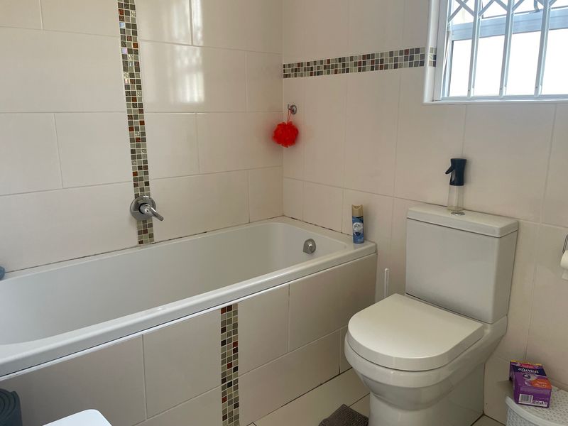 A functional bathroom featuring a bathtub with mosaic tile accents, a standard toilet, and tiled walls with security bars on the window.