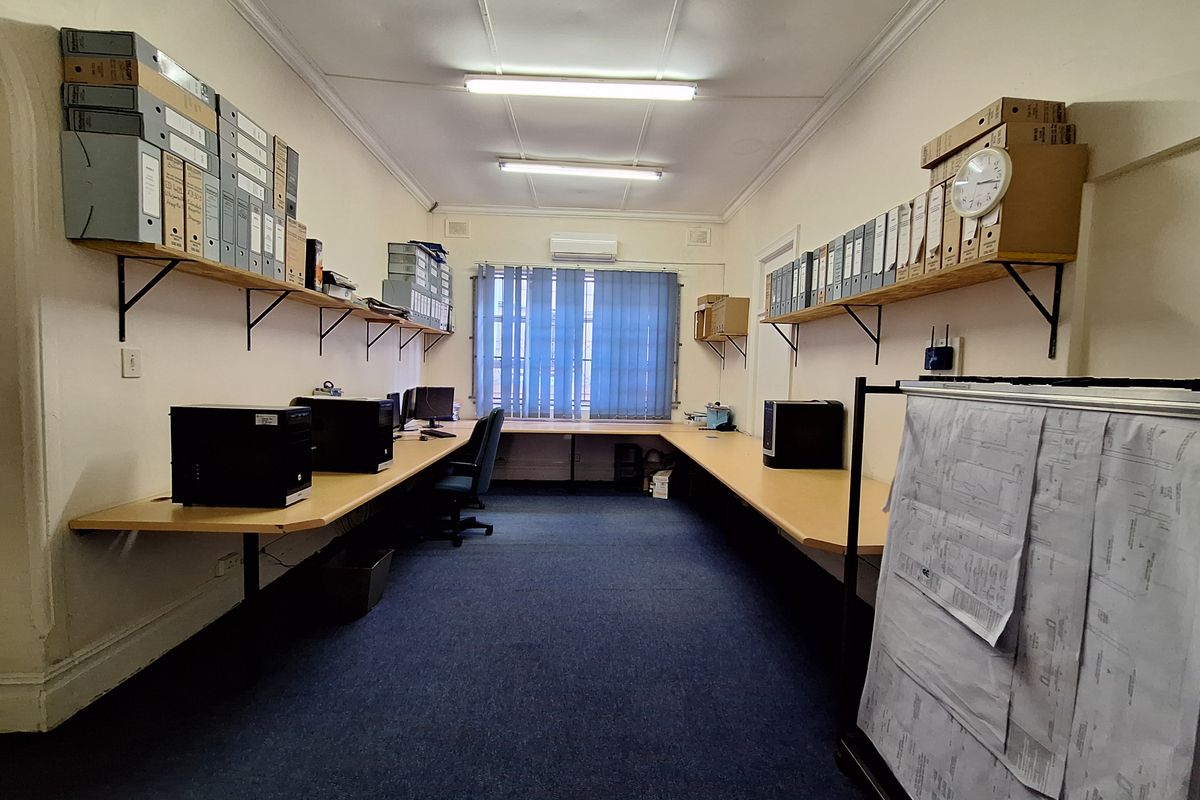A functional office space featuring built-in wrap-around desks, wall-mounted shelving units, and a wall-mounted air conditioning unit.