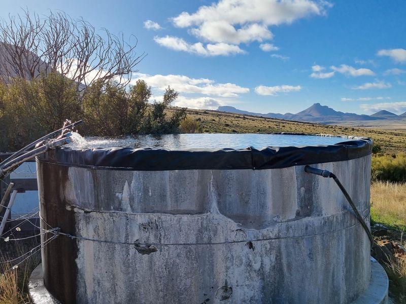 A circular concrete farm water reservoir or dam set against a scenic mountain landscape, featuring an active water inlet pipe.