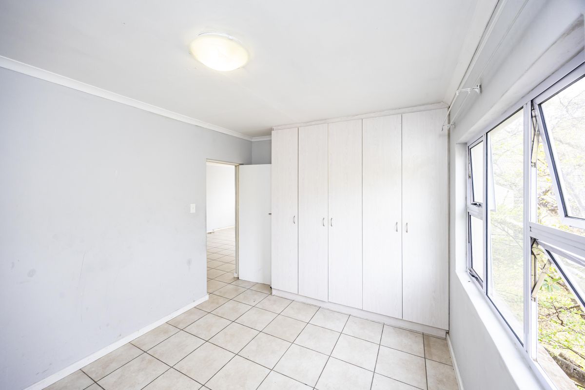 A bright, empty bedroom featuring neutral tiled flooring, built-in white cupboards, and large windows allowing for natural light.