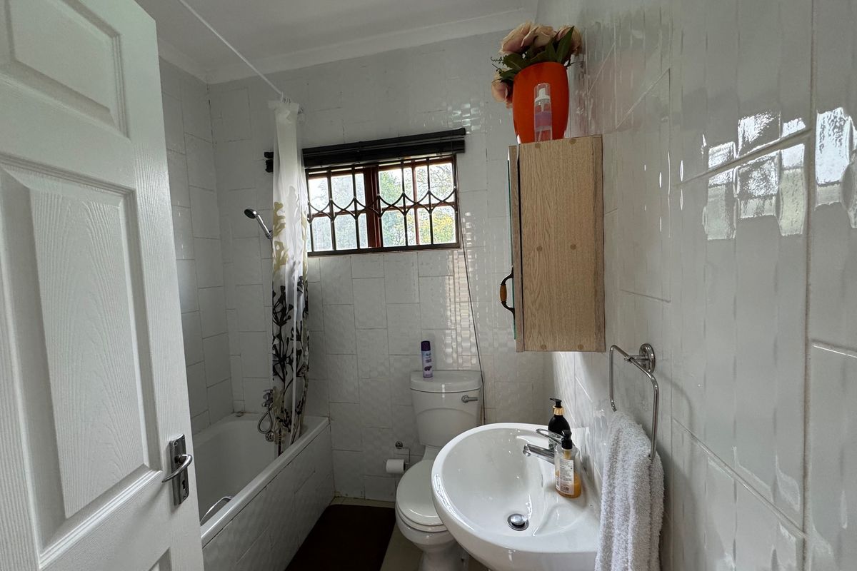Compact bathroom featuring a bathtub with shower, pedestal basin, and toilet, finished with white wall tiles and security bars on the window.