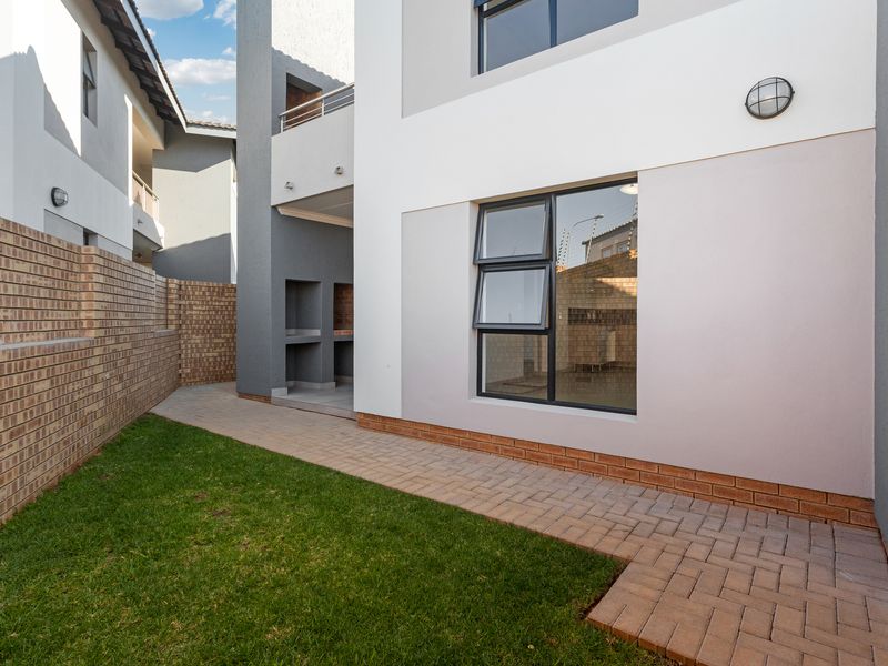 A well-maintained rear exterior area featuring a paved walkway, small lawn, brick boundary wall, and a built-in braai area.