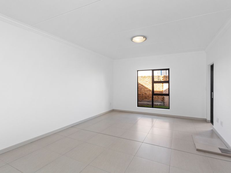 A bright, empty living area featuring neutral tiled flooring, clean white walls, and a modern black-framed window providing natural light.