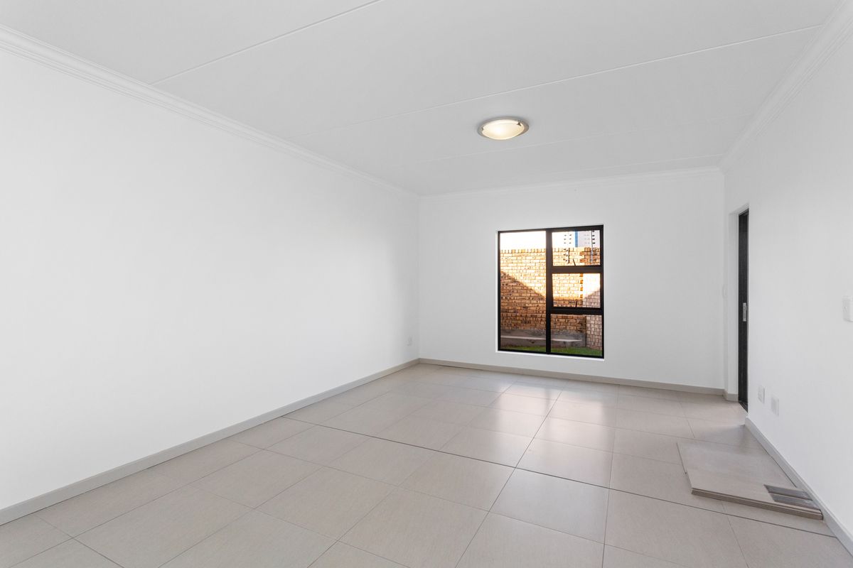 A bright, empty living area featuring neutral tiled flooring, clean white walls, and a modern black-framed window providing natural light.