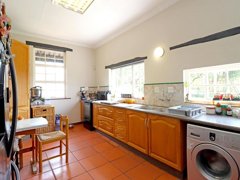 A functional kitchen featuring wooden cabinetry, laminate countertops, a double basin sink, and terracotta floor tiles with ample natural light from multiple windows.